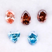 Five loose lab grown diamonds displayed on a white background, featuring three pink diamonds in oval, pear, and round cuts, alongside two vivid blue diamonds in heart and oval shapes, showcasing brilliant faceting and high clarity.