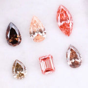 Six fancy colored lab grown diamonds including pear, marquise, and emerald cuts in pink, peach, champagne, and brown tones displayed on a white background, ideal for custom engagement rings and fine jewelry.