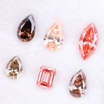 Six fancy colored lab grown diamonds including pear, marquise, and emerald cuts in pink, peach, champagne, and brown tones displayed on a white background, ideal for custom engagement rings and fine jewelry.