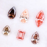 Six fancy colored lab grown diamonds including pear, marquise, and emerald cuts in pink, peach, champagne, and brown tones displayed on a white background, ideal for custom engagement rings and fine jewelry.