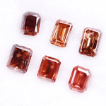 Six lab grown fancy orange pink emerald cut diamonds displayed on a white background, rectangular step cut loose grown diamonds in various reddish peach shades for engagement rings and fine jewelry.