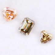 Three lab grown fancy color diamonds including pink heart cut, champagne emerald cut, and yellow heart shape loose diamonds for engagement ring and custom jewelry making