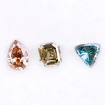 Three fancy color lab grown diamonds including pear cut pink diamond, asscher cut olive diamond, and trillion cut blue diamond displayed on white background for custom jewelry making