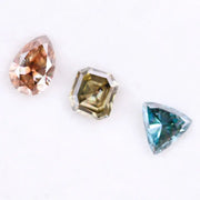 Three fancy color lab grown diamonds including pear cut pink diamond, asscher cut olive diamond, and trillion cut blue diamond displayed on white background for custom jewelry making