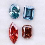 Four fancy color lab grown diamonds including oval and emerald cut blue stones and marquise and radiant cut red gemstones displayed on a white background.