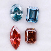 Four fancy color lab grown diamonds including oval and emerald cut blue stones and marquise and radiant cut red gemstones displayed on a white background.