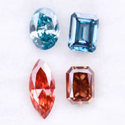 Four fancy color lab grown diamonds including oval and emerald cut blue stones and marquise and radiant cut red gemstones displayed on a white background.