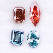 Four fancy color lab grown diamonds including oval and emerald cut blue stones and marquise and radiant cut red gemstones displayed on a white background.