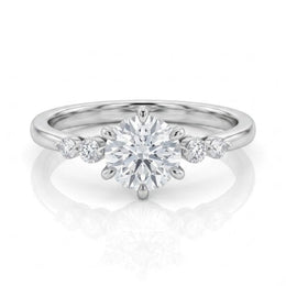 A silver solitaire engagement ring with a round-cut center stone and smaller accent stones on the band, shown on a clean white background with a soft reflection.