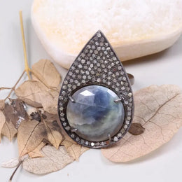 Teardrop-shaped jewelry piece with a blue gemstone on a white surface with leaves.
